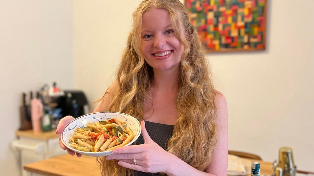 blonde woman holding a pasta dish and smiling in her kitchen